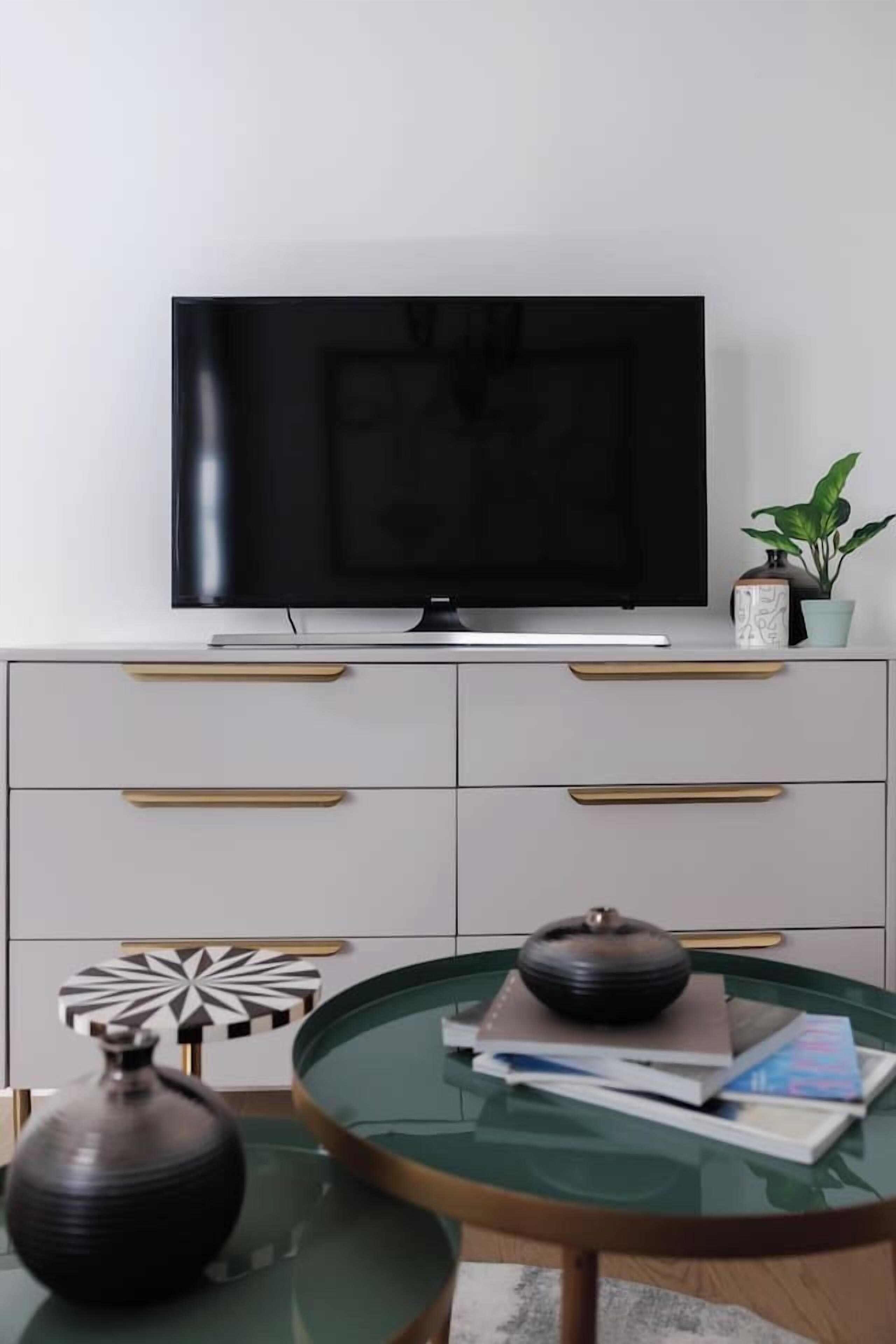 A modern living room with a flat-screen TV, a white cabinet with gold handles, a glass coffee table with books and a decorative bowl, and a potted plant.