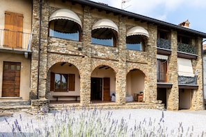 A stone building with multiple arches, balconies, and a gravel driveway with lavender plants. - Cottage Zia Cherubina (Colle Brianza)