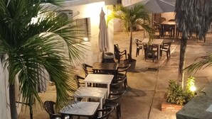 Un espace extérieur aménagé avec des tables et des chaises, entouré de plantes en pot et surmonté d’un parasol en chaume.