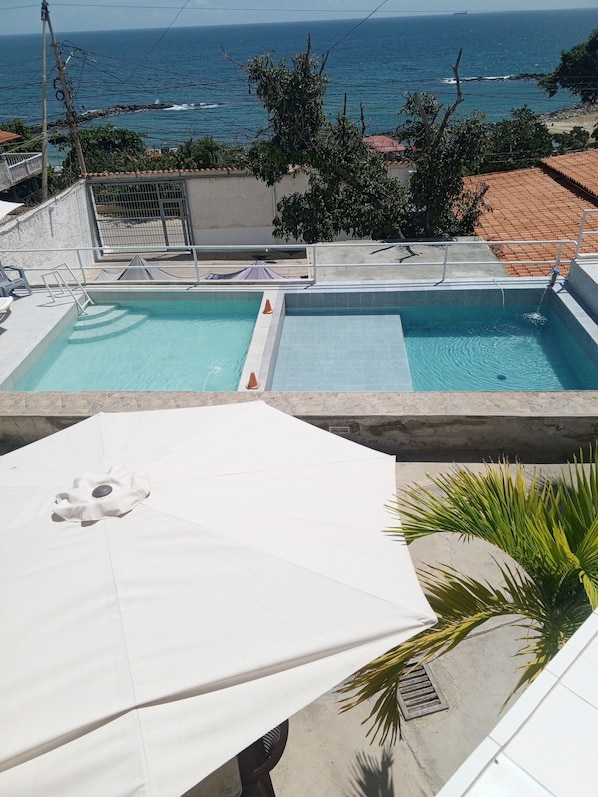 Piscina com vista para o oceano, rodeada por um deck de concreto e um guarda-sol branco.