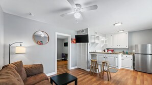 Un espacio habitable compacto con sofá, una mesita, una zona de cocina con armarios blancos, refrigerador y ventilador de techo.