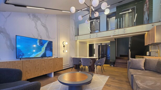 A modern living room with a flat-screen TV, wooden furniture, a dining area, and a staircase leading to an upper level. - Apartments Lura (Peć)