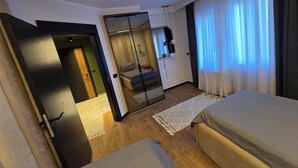 A modern hotel room with a large bed, a smaller bed, a walk-in closet, and a view of the outdoors through sheer curtains. - Apartments Lura (Peć)