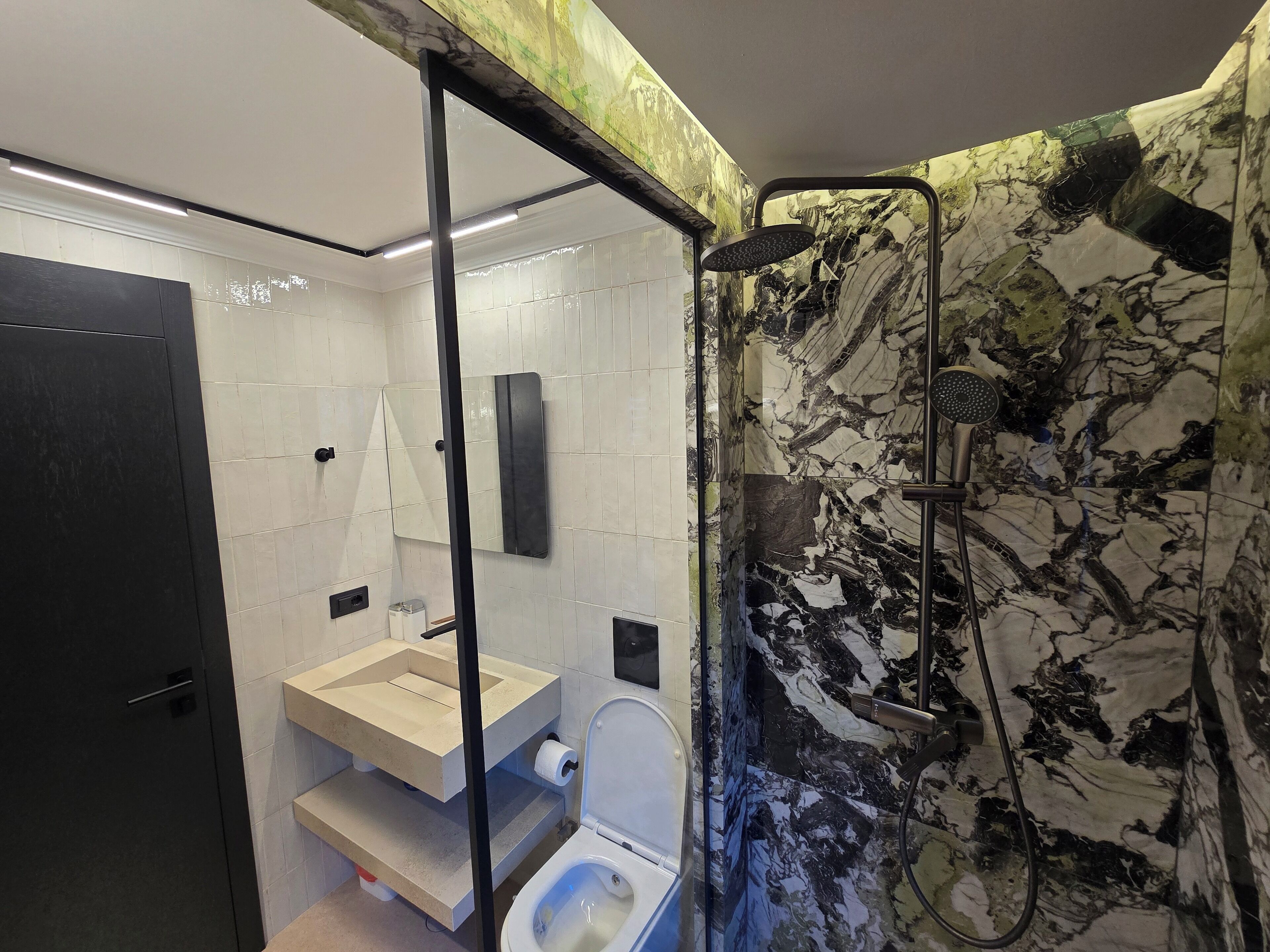 Ein modernes Badezimmer mit Duschwand aus Marmor, Toilette und Waschbecken mit Spiegel darüber.