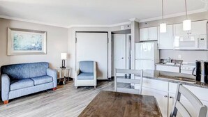 Une chambre d’hôtel moderne équipée d’un canapé, d’un fauteuil, d’une table à manger et d’un coin cuisine avec un four à micro-ondes et une cuisinière, ainsi qu’un tableau encadré au mur.