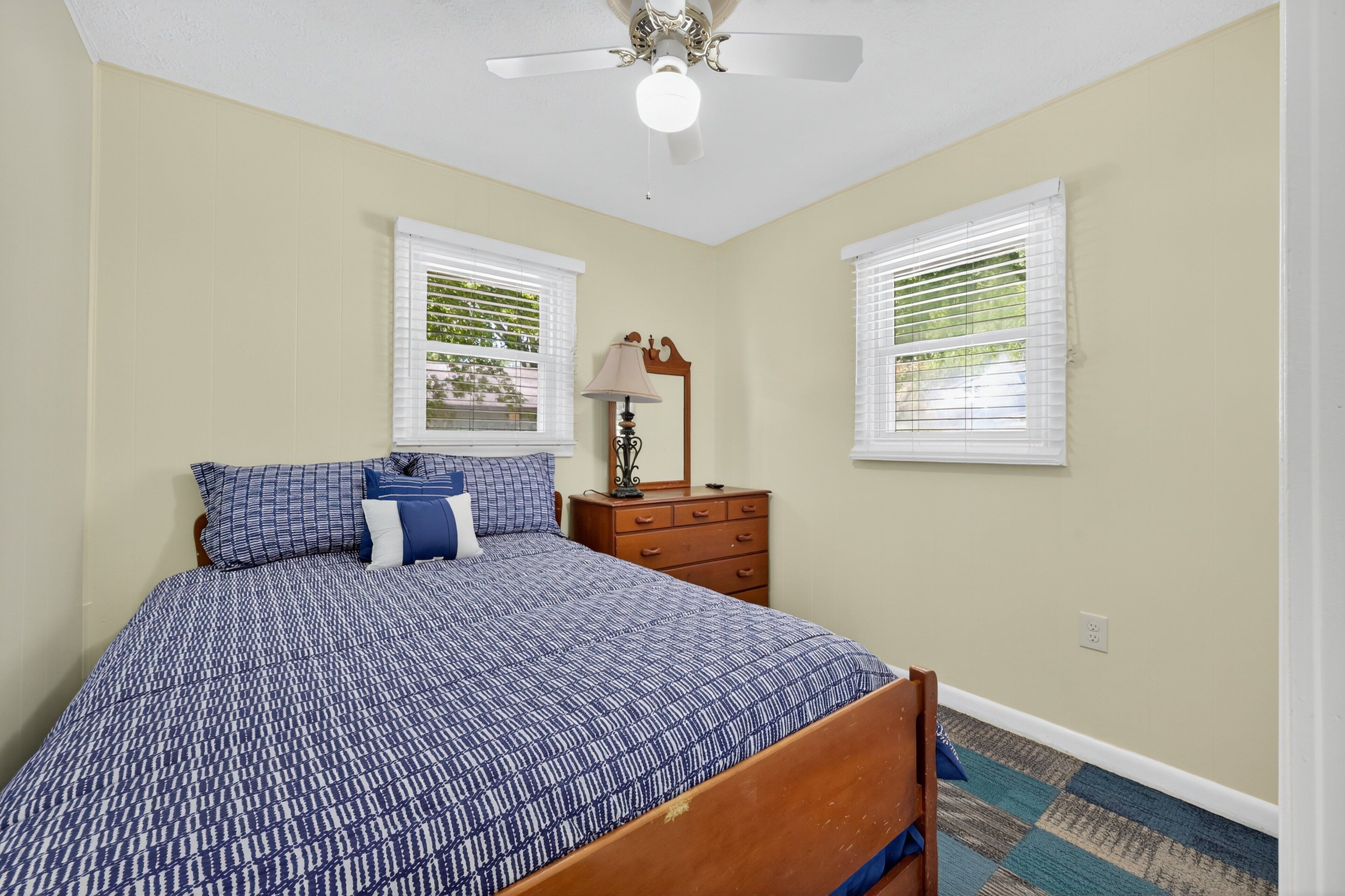 A bedroom with a bed, dresser, two windows with blinds, a ceiling fan, and a lamp.