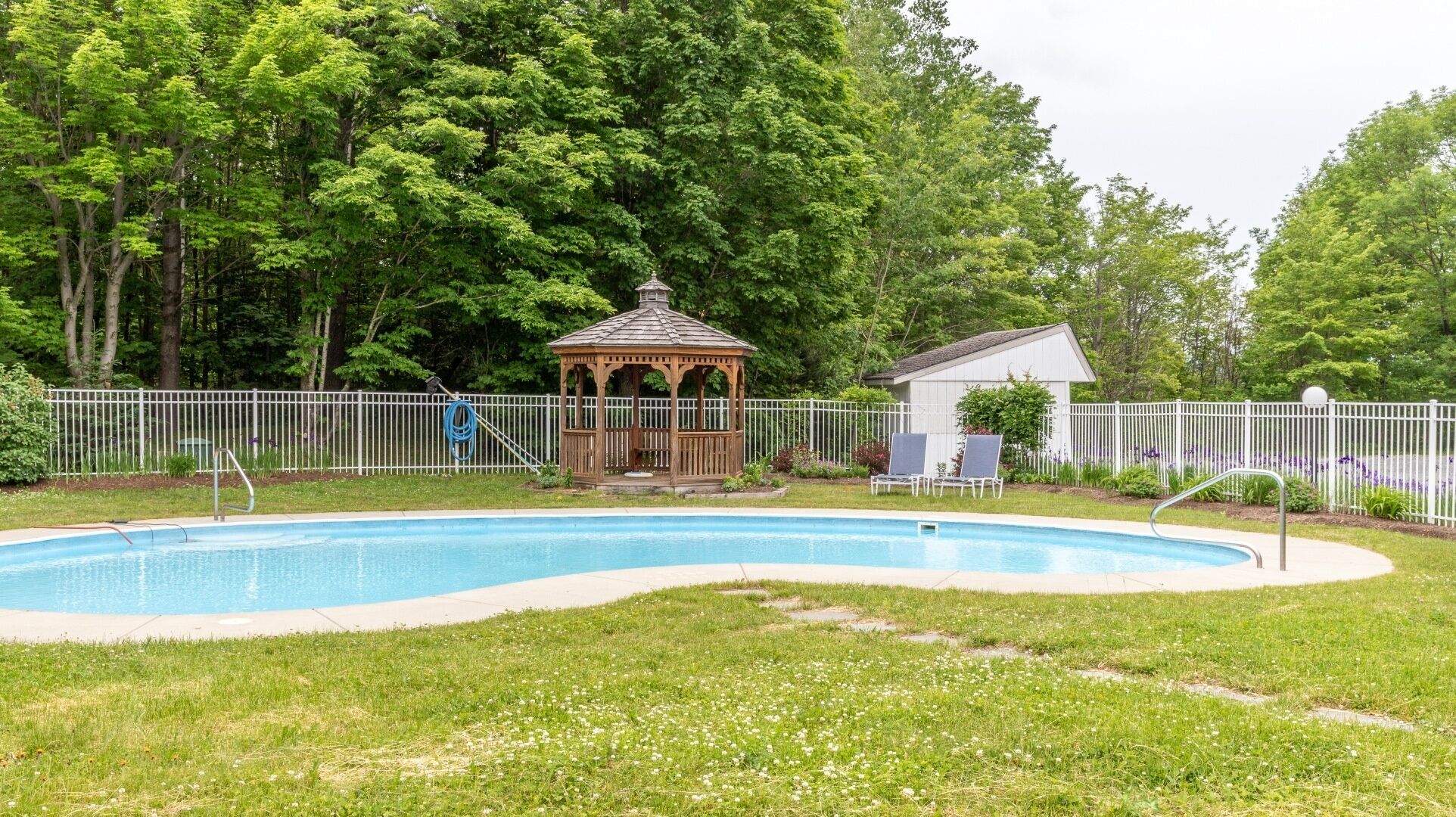 A swimming pool with a gazebo and lounge chairs, surrounded by a white fence and lush greenery.
