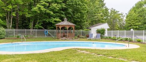 A swimming pool with a gazebo and lounge chairs, surrounded by a white fence and lush greenery.
