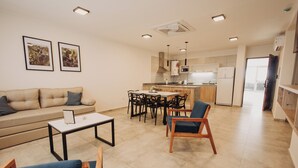 A modern living room with a sofa, coffee table, and chairs, a kitchen with a dining area, and a hallway leading to a door. - Edificio Coirón Premium (San Rafael)