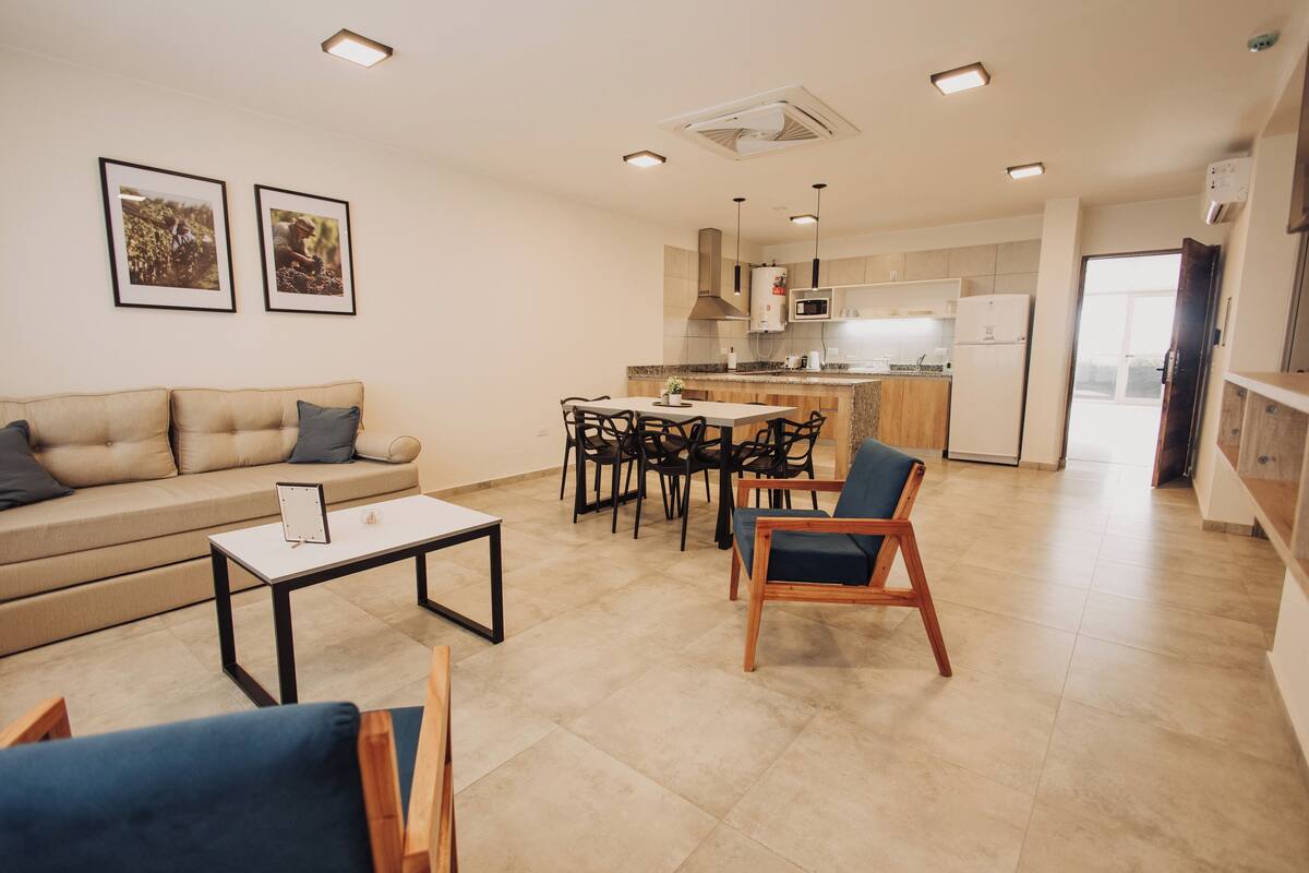 A modern living room with a sofa, coffee table, and chairs, a kitchen with a dining area, and a hallway leading to a door.