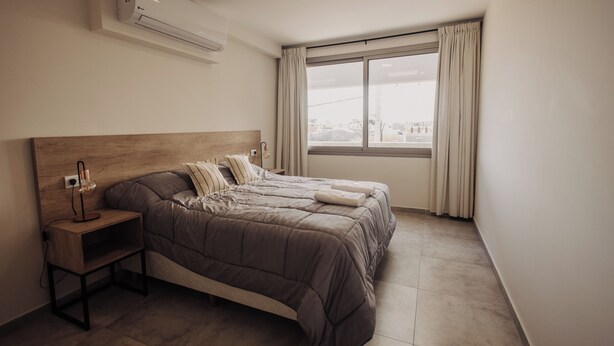 A bedroom with a wooden headboard, a bed with a brown quilt, a bedside table with a lamp, and a window with curtains. - Edificio Coirón Premium (San Rafael)