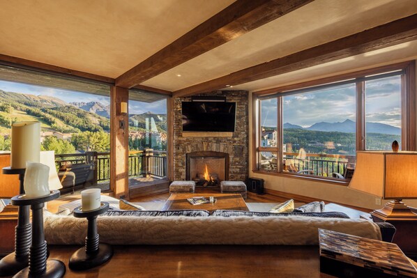 A spacious living room with a stone fireplace, a flat-screen TV, and a large window offering a mountain view.
