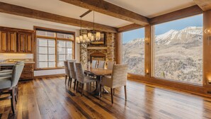 A spacious dining area with a large wooden table and chairs, a stone fireplace, and a view of mountains through large windows. - See Forever Home by InvitedHome | Walk to Ski, Ski Valet, Large Deck with Views (Telluride)