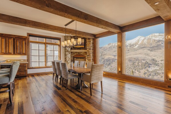 A spacious dining area with a large wooden table and chairs, a stone fireplace, and a view of mountains through large windows.