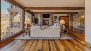 A spacious living room with a fireplace, wooden beams, stone walls, and large windows. - See Forever Home by InvitedHome | Walk to Ski, Ski Valet, Large Deck with Views (Telluride)