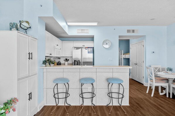 A modern kitchen with white cabinets, a central island with stools, a clock on the wall, and a dining area with a table and chairs. - Celadon condo - beach access, pools and hot tub (Panama City Beach)