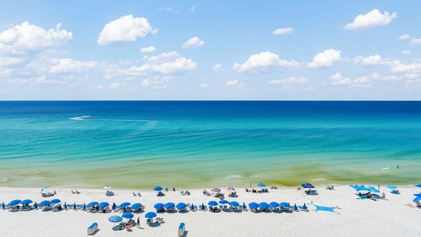 A beach with white sand and clear turquoise water. There are blue umbrellas and lounge chairs along the shore. - Celadon condo - beach access, pools and hot tub (Panama City Beach)