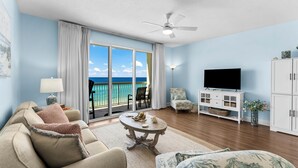 A living room with a large window, a ceiling fan, a TV, and a white cabinet. - Celadon condo - beach access, pools and hot tub (Panama City Beach)