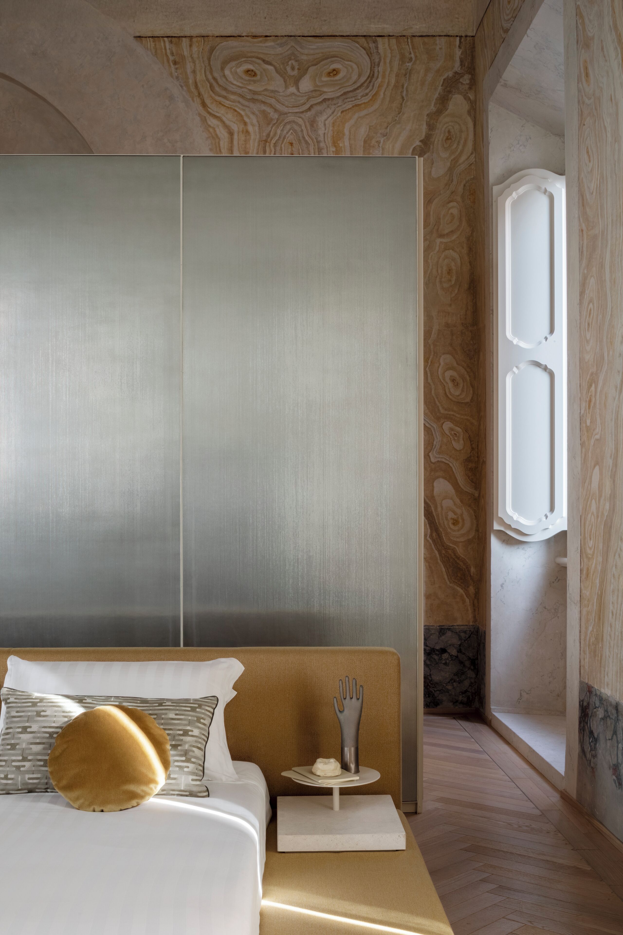 Une chambre d’hôtel moderne avec un grand lit, une tête de lit en bois, une petite table agrémentée d’un objet décoratif, et une vue sur un mur de marbre et une porte.