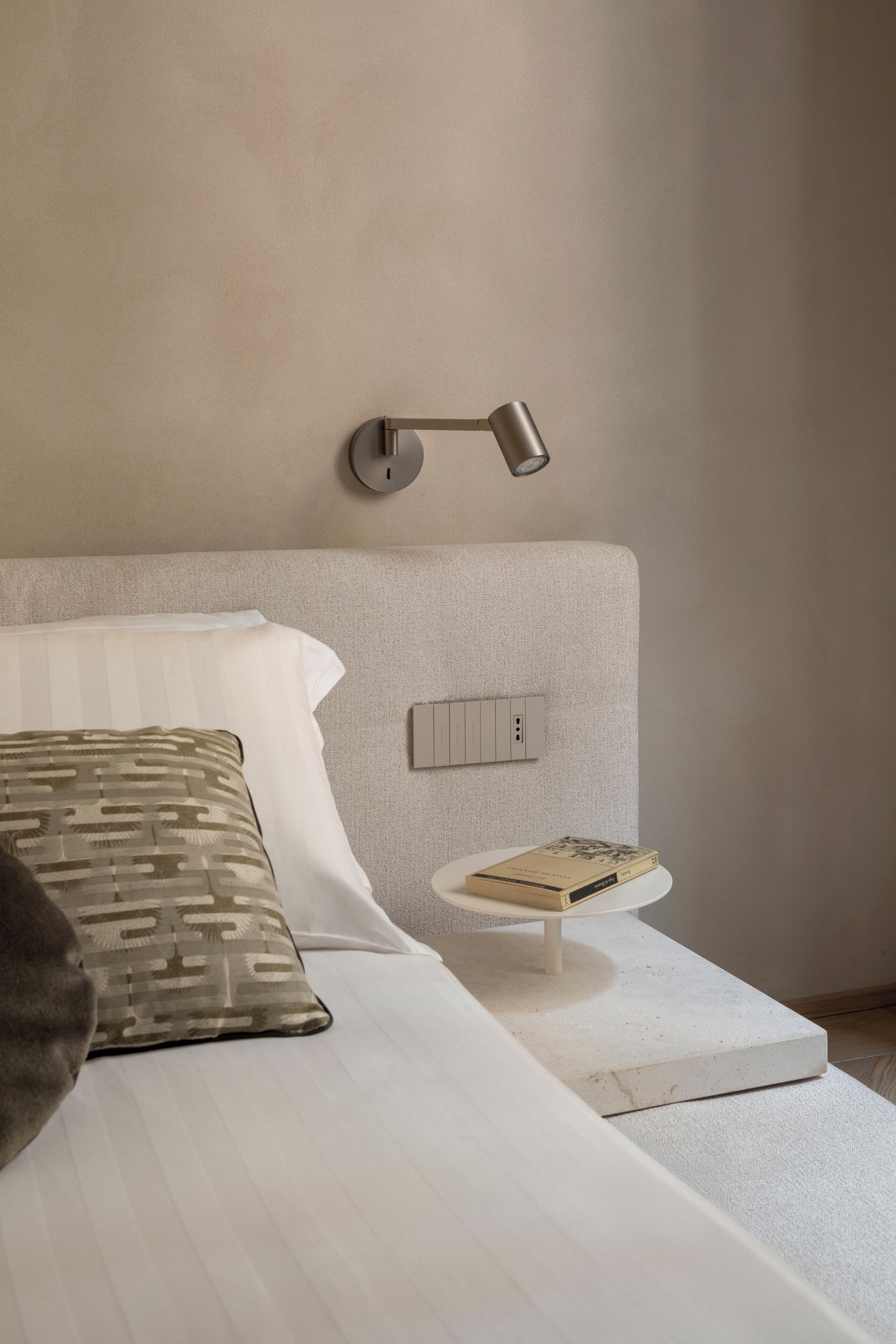 A neatly made bed with a patterned pillow, a bedside table with a book, and a wall-mounted light fixture.