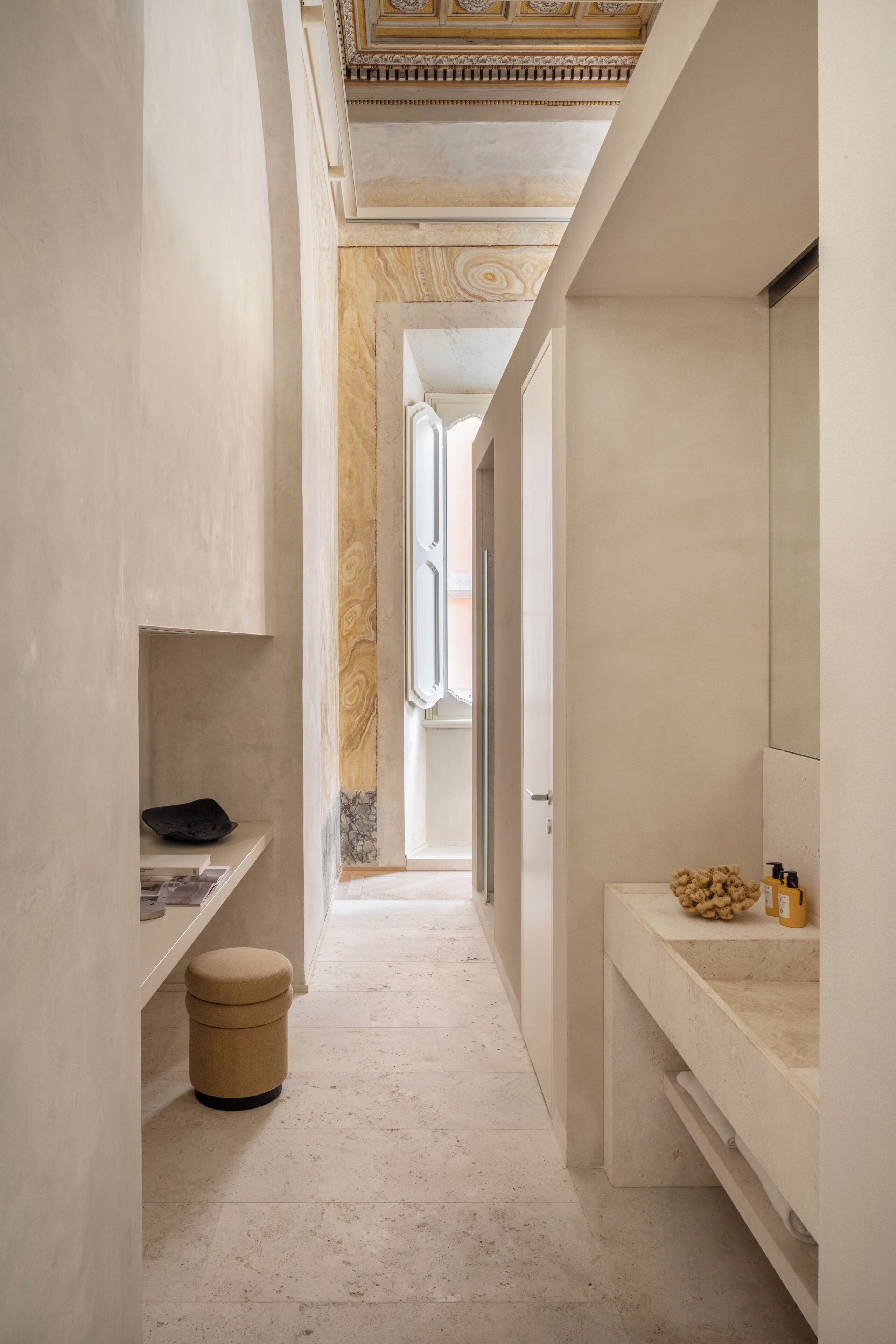 Un pasillo estrecho con piso de mármol, un cubo de basura pequeño, un banco blanco y un espejo en la pared derecha.