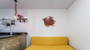 A small, compact room with a yellow sofa, a kitchen area with utensils and a hanging light, and a decorative wall piece. - Loft em Condom nio com mar em Mangaratiba (Mangaratiba)
