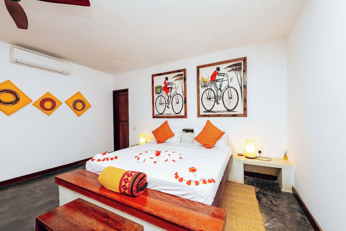 A hotel room with a bed, a bench, a nightstand, wall art, and an air conditioning unit.