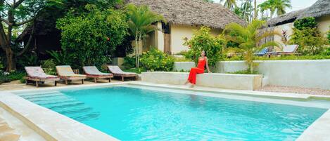 A swimming pool with lounge chairs, a person in red, and a thatched-roof building.
