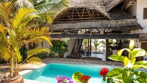 A pool area with a thatched-roof structure, palm trees, and a staircase leading to an upper level. - Hekaya Zanzibar (Michamvi)