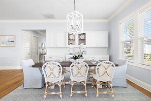 Uma área de refeições com mesa de madeira, cadeiras de vime branco, um lustre branco e vista para o exterior através de uma janela com persianas.
