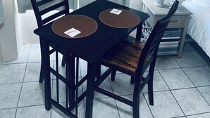 Une petite pièce, bien rangée, avec une table et des chaises, une table de chevet avec une lampe et un tableau encadré au mur.