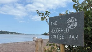 Skilt for The Woodshed Coffee Bar med udsigt til en strand og en båd i baggrunden.