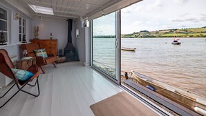 Et værelse med et stort vindue der kigger ud over en sø, en pejs og et siddeområde med en stol og et bord.