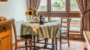 A dining area with a table set for two, a wooden chair, a radiator, and a window with curtains. - Poplar Cottage | Sleeps 3 | Near Broadway (Dumbleton)