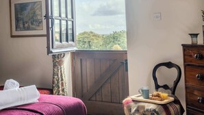 A room with a bed, a wooden gate, a chair with a tray of breakfast, and a painting on the wall. - Poplar Cottage | Sleeps 3 | Near Broadway (Dumbleton)