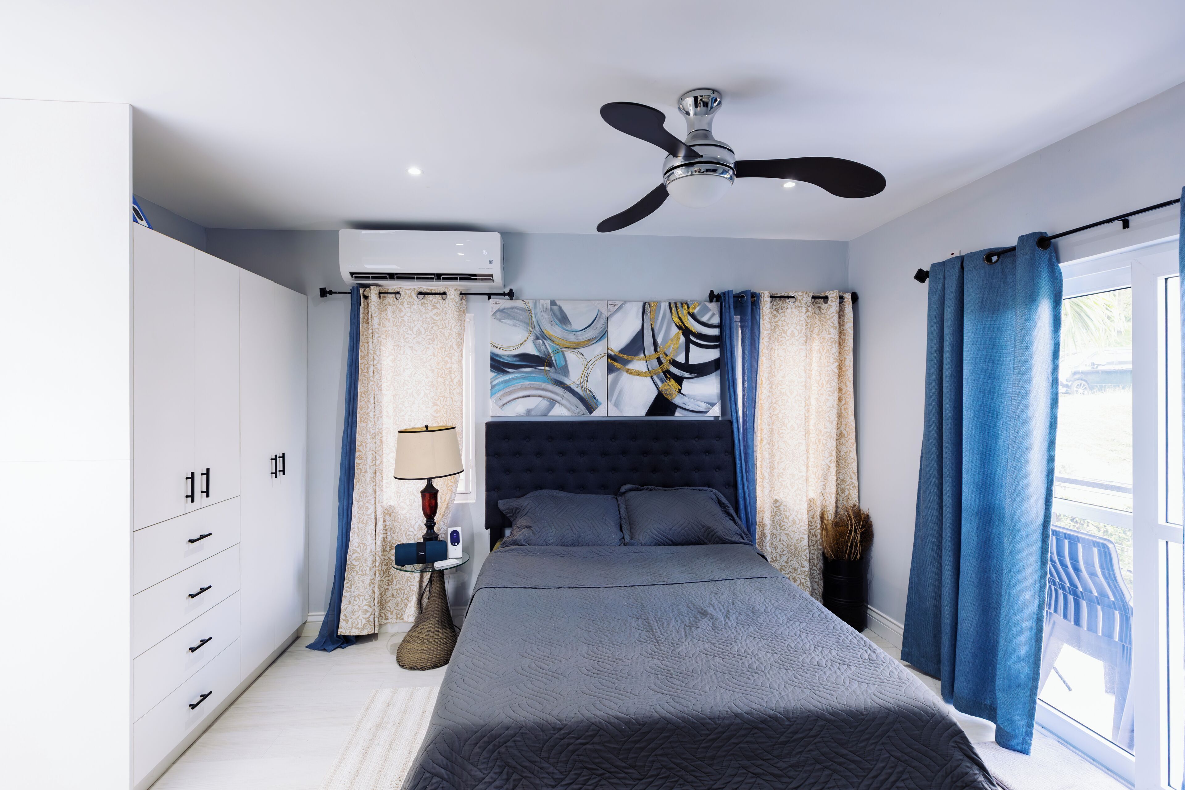 Un dormitorio con cama, ventilador de techo, aire acondicionado, mesita de noche con lámpara y ventana con cortinas.