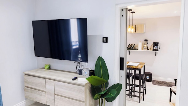 A modern living room with a flat-screen TV, a wooden cabinet, a large green plant, and a view into the dining area with a kitchen visible. - Cozy 1-bedroom apartment in beautiful Grande Riviere with AC (Grande Riviere)
