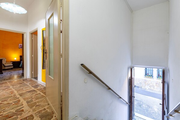 Un couloir avec un sol en pierre, une porte donnant sur une pièce dont un mur est jaune, et une fenêtre donnant sur la rue.