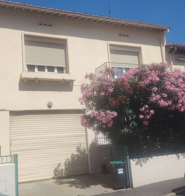 Un bâtiment à deux étages doté d’une porte de garage, d’un balcon avec une rambarde en métal et d’un grand arbuste en fleurs devant.