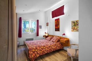 A bedroom with a bed, a nightstand, a chair, a window, and artwork on the walls. - Pech Chabrol: Family Cottage with Pool and Vineyard View (Ribagnac)