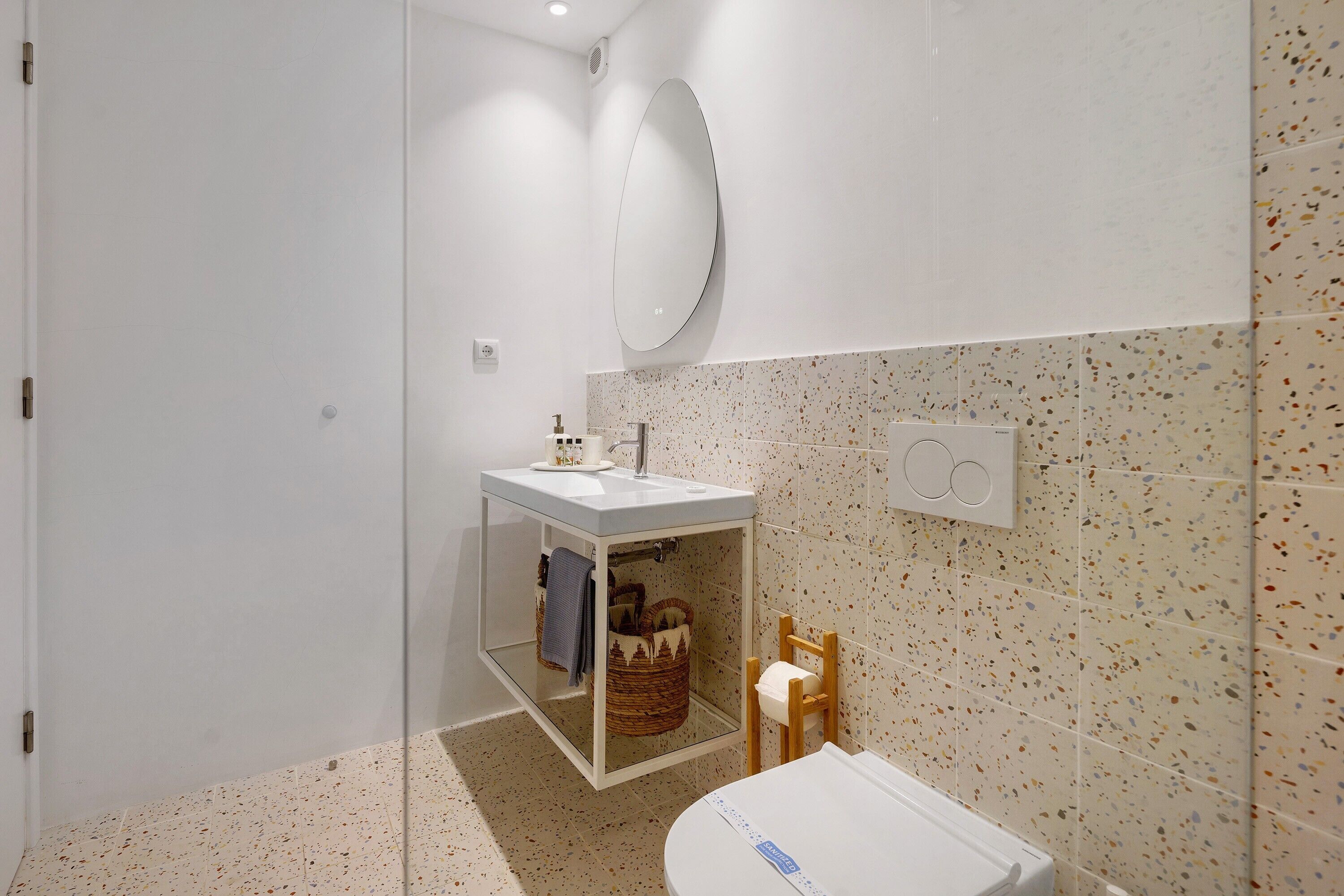 Une salle de bain avec un lavabo blanc, un miroir, un porte-serviettes, une petite chaise en bois et un panier décoratif.