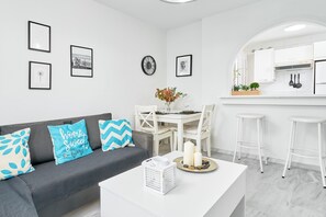 A modern kitchen with white cabinets, a dining area with a white table and chairs, and a living area with a grey sofa and decorative pillows.