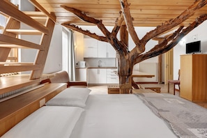 A bedroom with a large bed, wooden headboard, a wooden tree trunk centerpiece, a view of a kitchen, and a television on a cabinet.