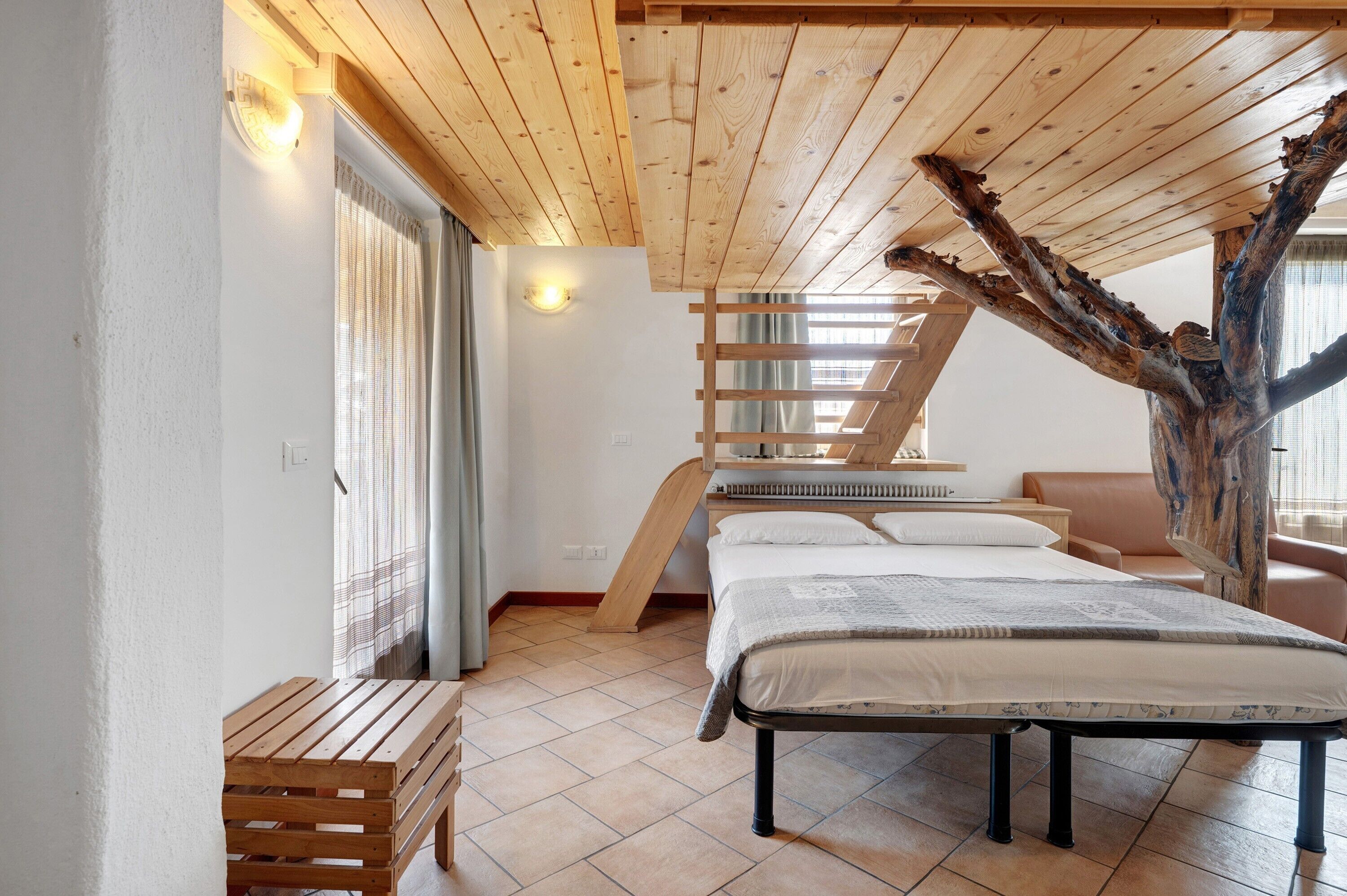 Una camera da letto con soffitto spiovente, una panca in legno, un letto e un’imponente ramo d’albero utilizzato come elemento architettonico originale.