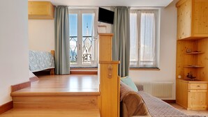 A room with a wooden staircase, a bed with patterned bedding, a wooden cabinet, and a window with a view of a tower. - Room 'Vento Alpino Campanile' with Balcony and Wi-Fi (Coredo)