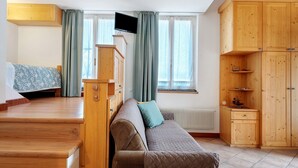 A cozy room with a sofa, a television, wooden cabinets, and a window with curtains. - Room 'Vento Alpino Campanile' with Balcony and Wi-Fi (Coredo)