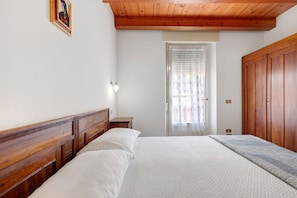 Un dormitorio con cabecera de madera, cama con ropa blanca, mesita de noche, armario de madera y una puerta con ventanal.