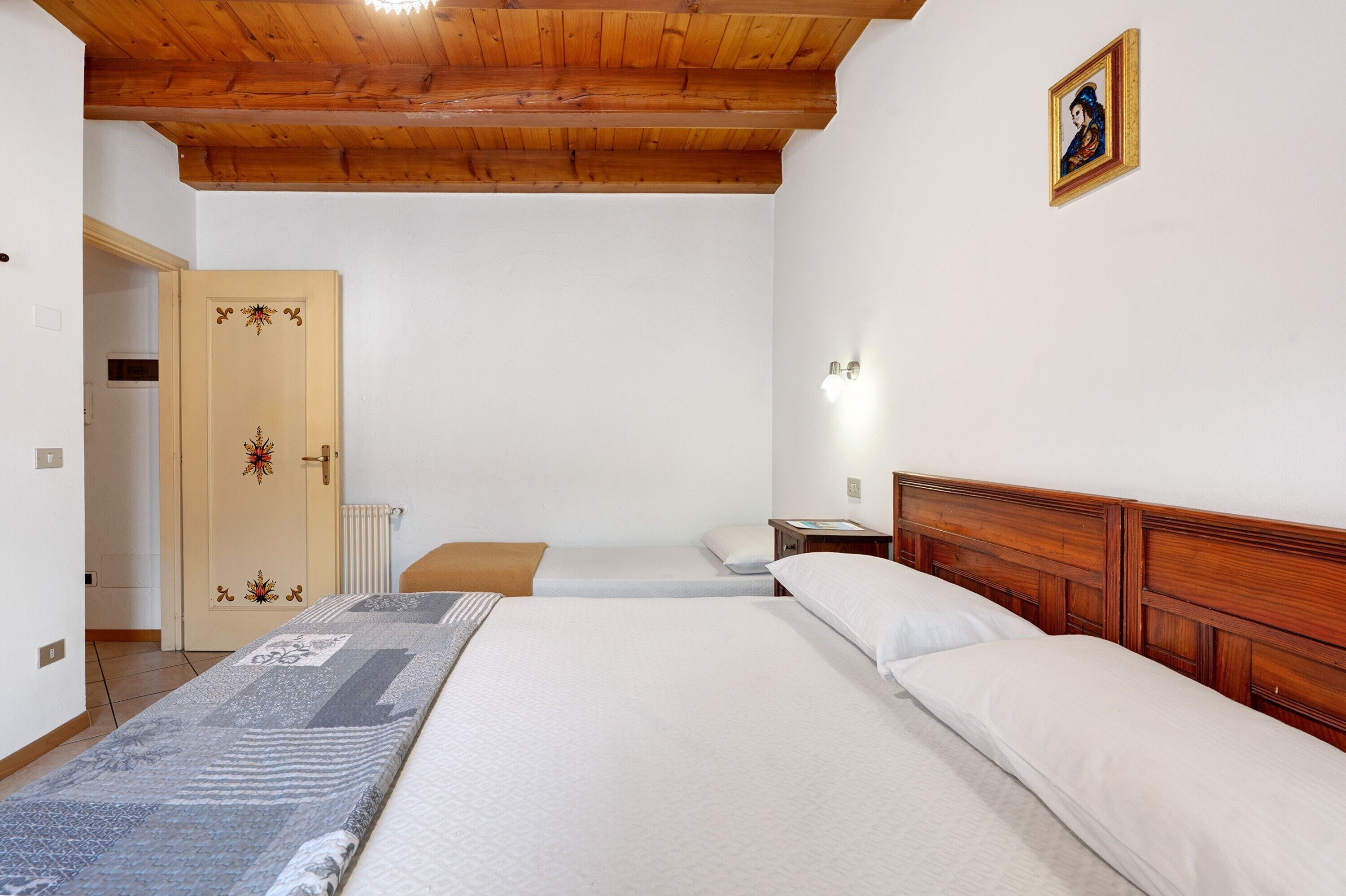 Ein Zimmer mit zwei Betten, Holz-Kopfteilen, einer Holzbalkendecke, einer weißen Tür mit Verzierungen und einem gerahmten Bild an der Wand.