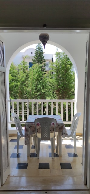 Un espace extérieur abrité, aménagé pour les repas, avec une table et des chaises, une rambarde blanche, et une vue sur la verdure et les bâtiments en arrière-plan.