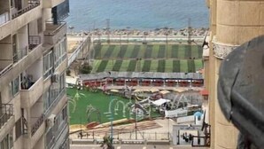 A high-rise building overlooking a coastal area with a sports field and parked cars. - Sea view in gleem luxury apartments - Saba Basha - alexandria (Alexandria)
